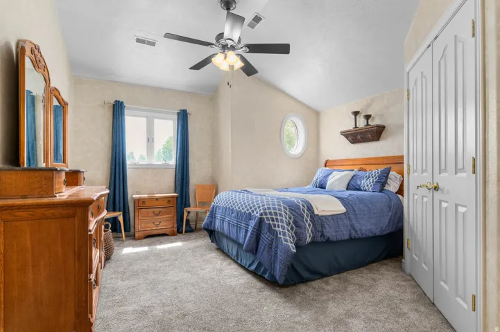 Carpeted bedroom with a vaulted ceiling, a ceiling fan with integrated lighting, a double-hung window, an oval window, and bi-fold closet doors
