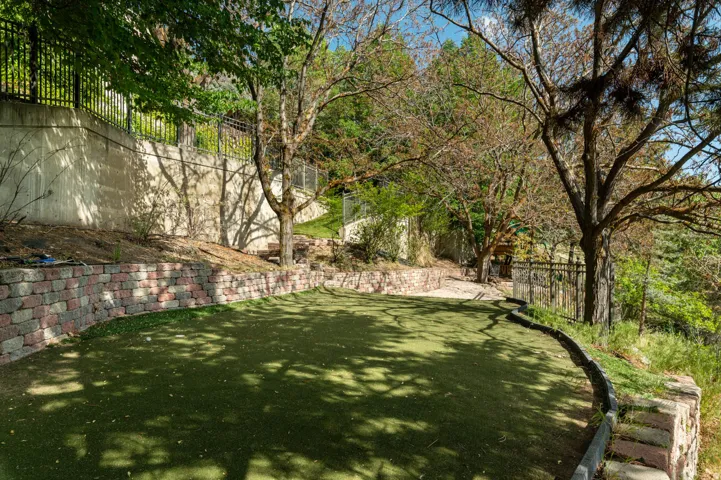 Artificial turf lawn with two-tone retaining wall, mature trees, and a concrete retaining wall with black metal fencing