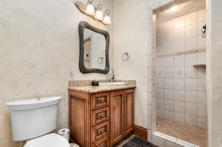 Bathroom featuring a single vanity with wood cabinetry and a graite countertop, an ornate framed mirror, a three-light vanity fixture, and a tiled shower stall with a built-in shelf