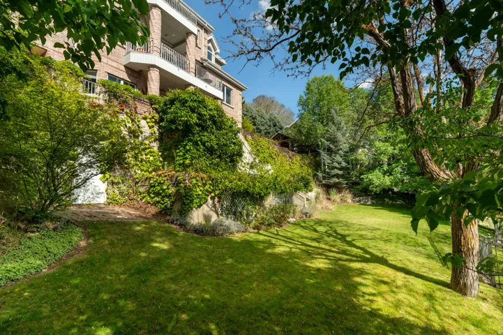 Expansive lawn bordered by mature trees and lush landscaping, with a multi-level brick exterior featuring a balcony