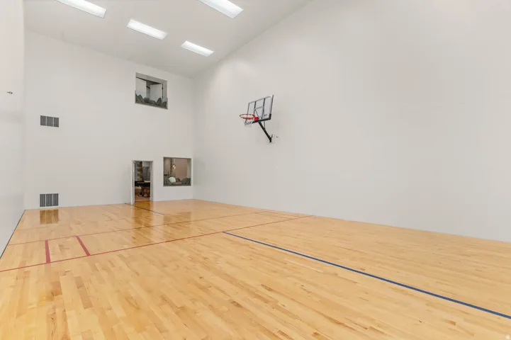 Indoor sports court featuring hardwood flooring with court markings, a wall-mounted basketball hoop, high ceilings, and recessed rectangular lighting