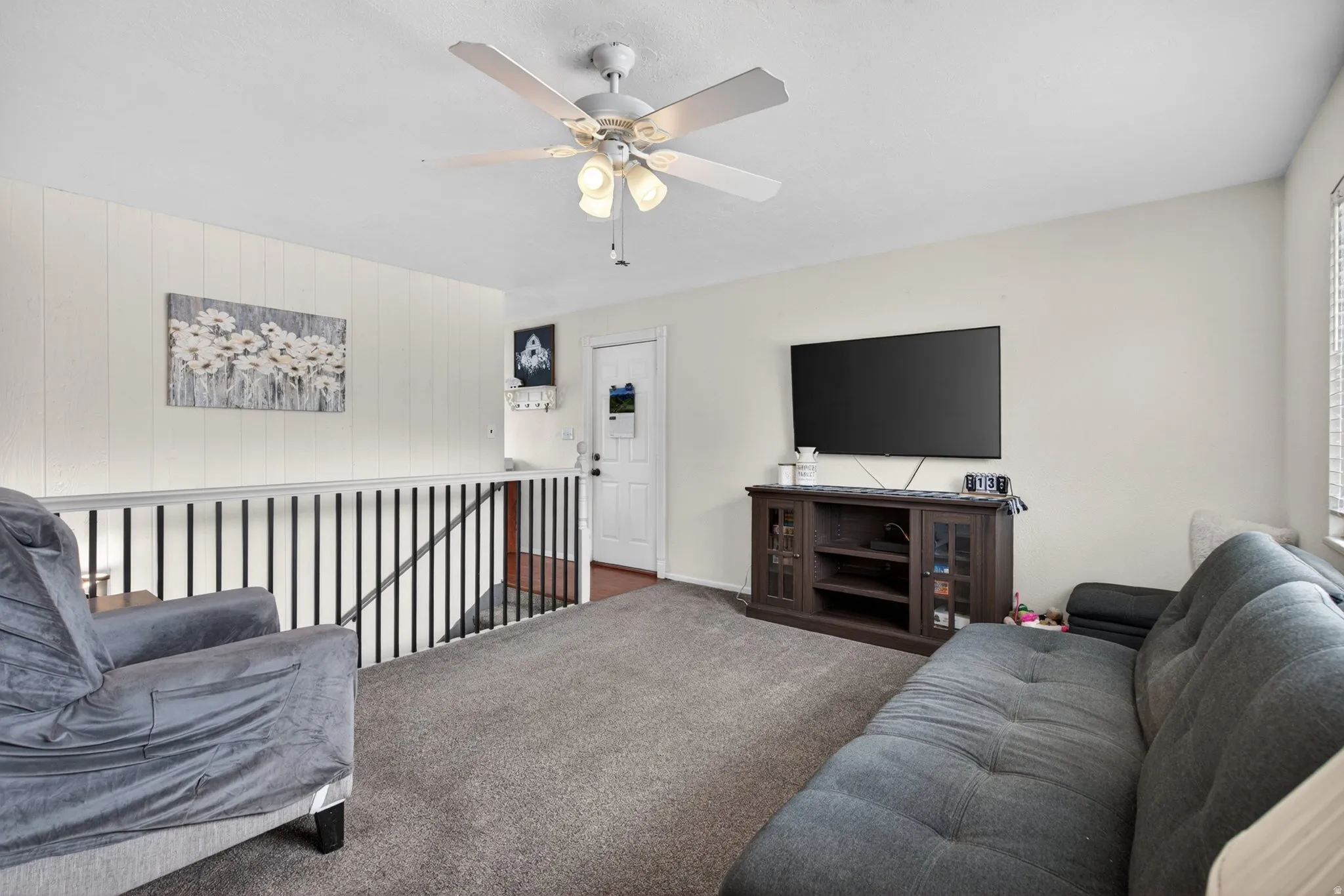 Carpeted upper-level landing featuring a white ceiling fan with integrated lighting, white paneled wall accents, a white railing, and a window with horizontal blinds