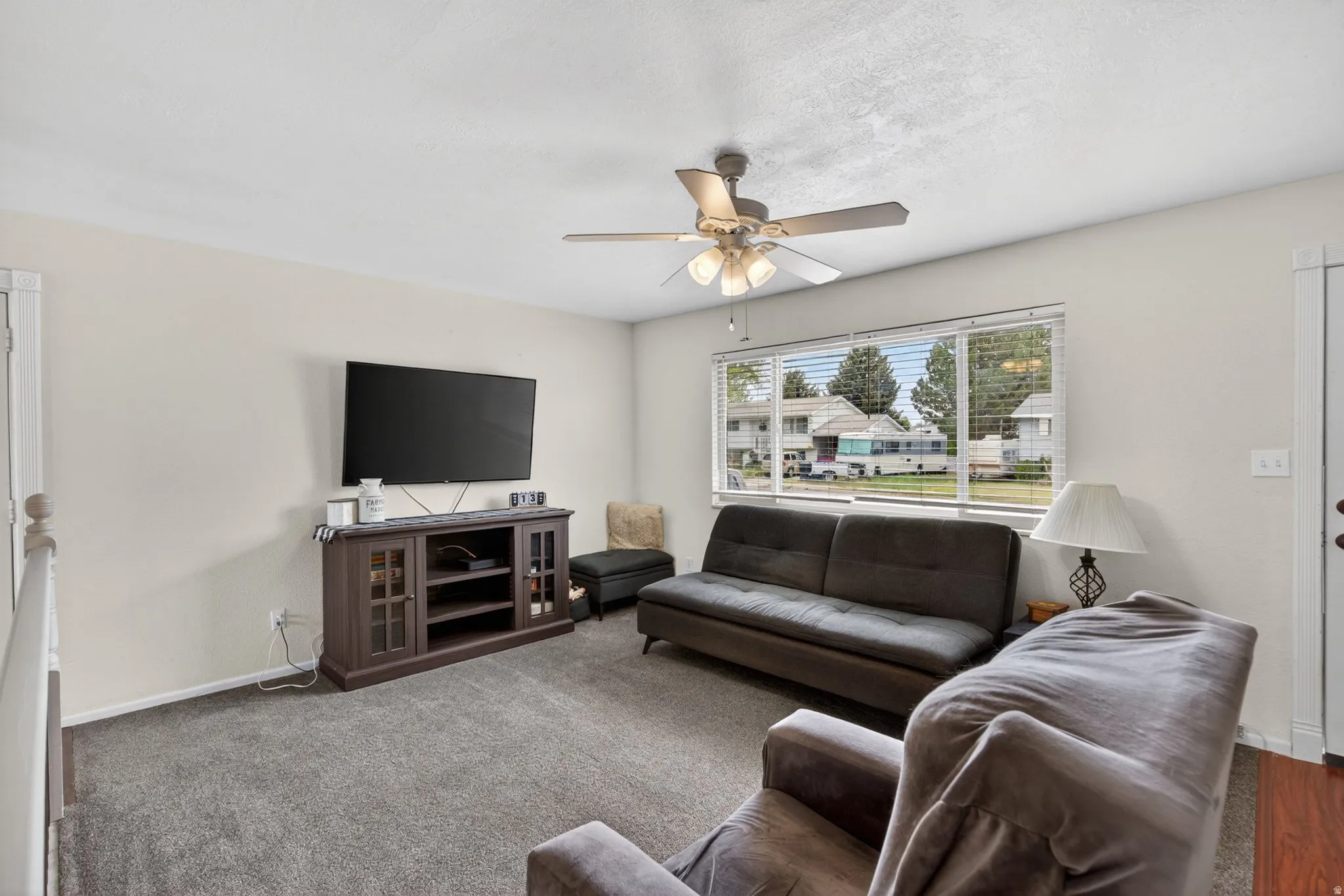 Living area featuring light neutral wall paint, dark grey carpet, a ceiling fan with integrated lighting, and a large window with horizontal blinds