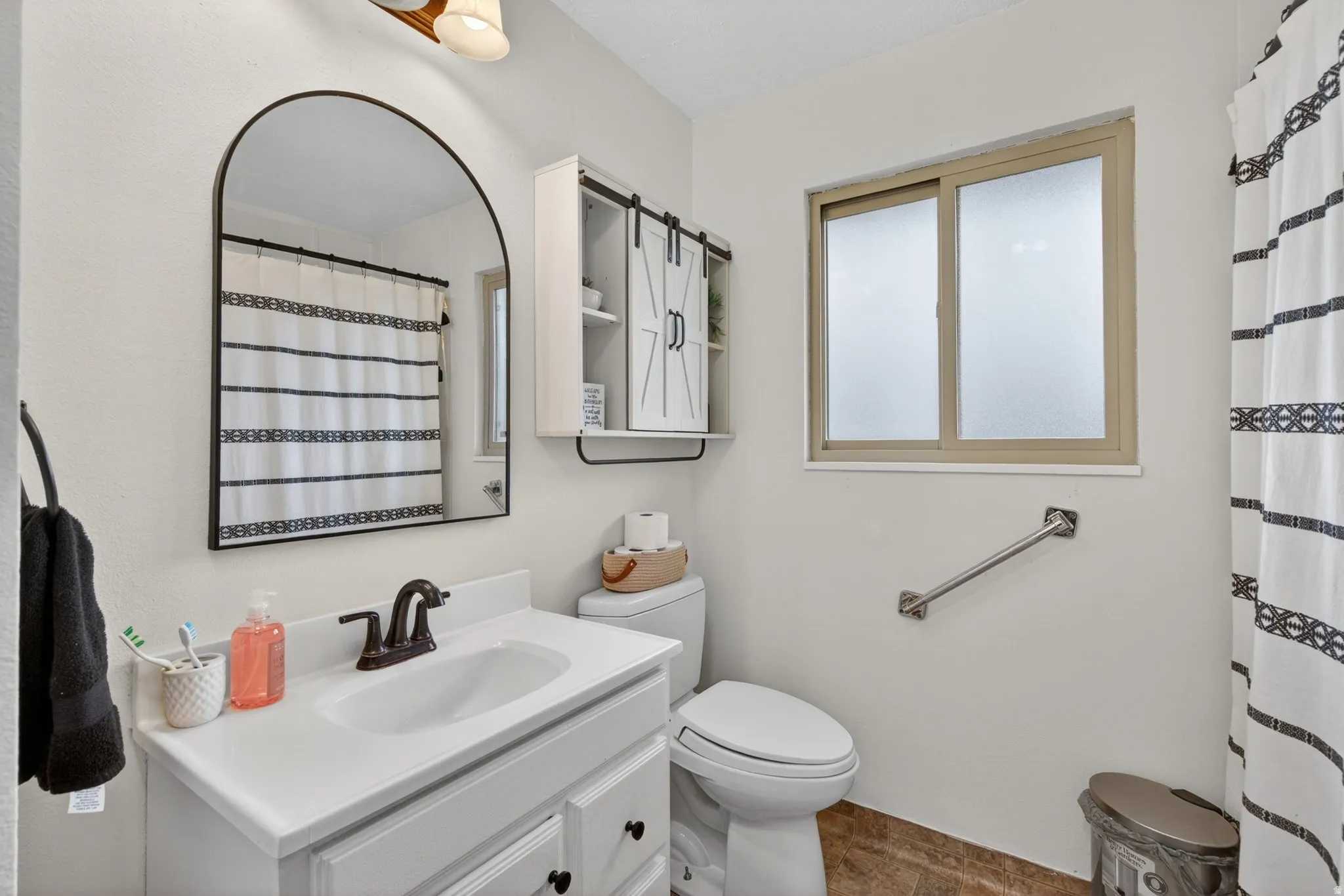 Bathroom featuring a single vanity with white countertop, arched mirror, sliding window with frosted panes, and a wall-mounted grab bar