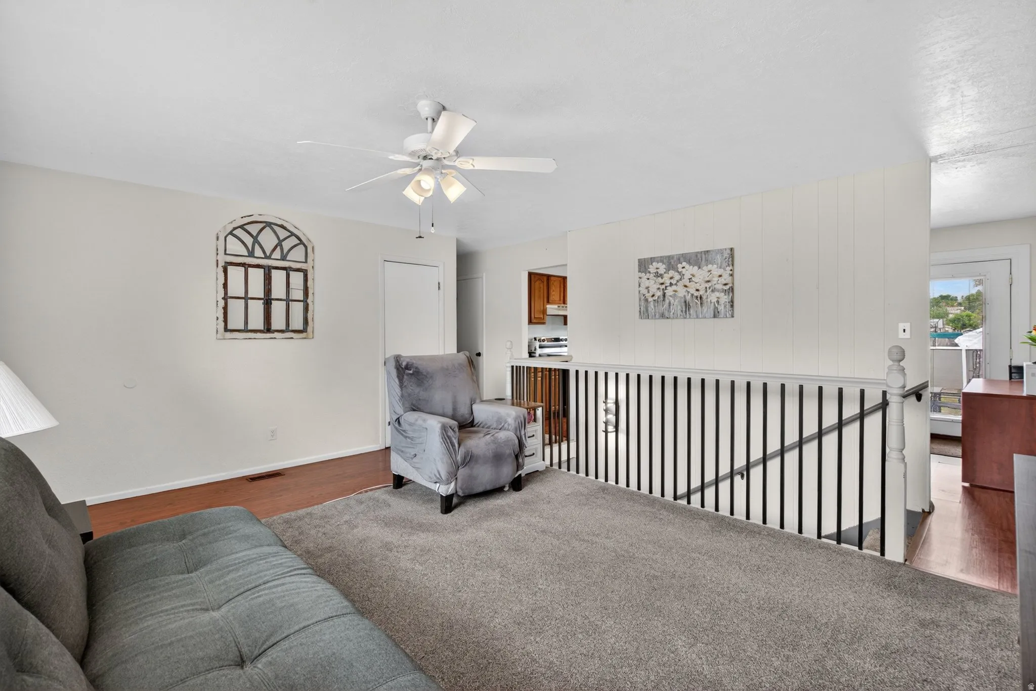 Carpeted living area featuring a ceiling fan with integrated lighting, wood-finish flooring, a white spindle railing, and a partial view of a kitchen with wood cabinetry