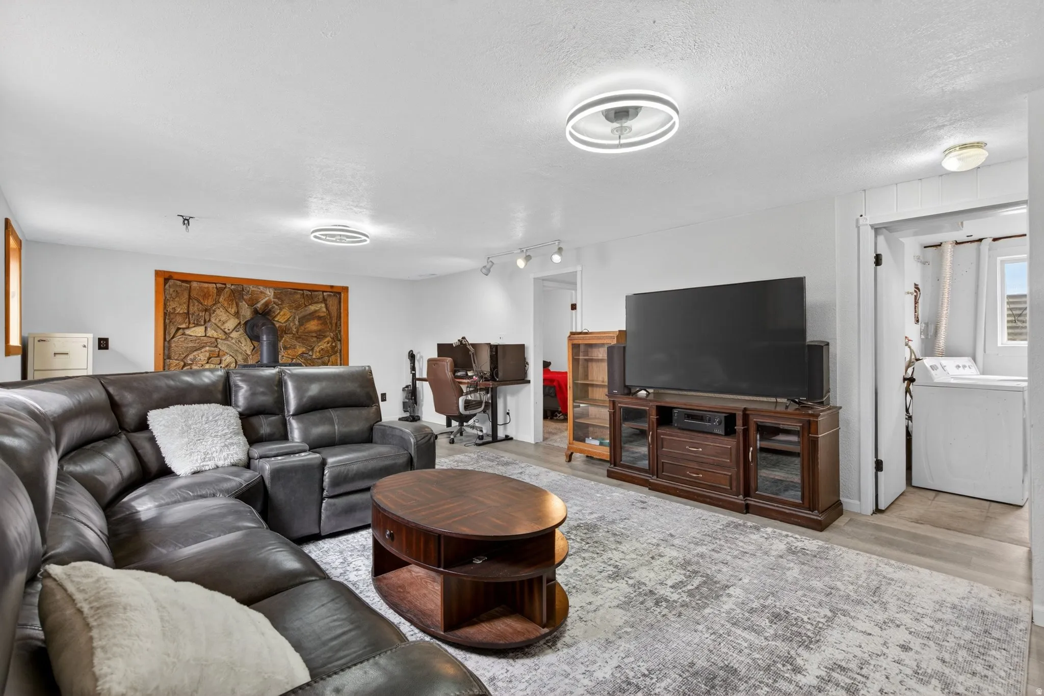 Spacious living area featuring a stone accent wall with wood trim, light wood-finish flooring, white walls, and contemporary ceiling light fixtures