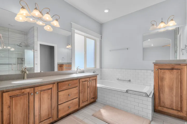 Bathroom featuring a dual vanity with light-toned countertops and wood-finish cabinetry, a large soaking tub with subway tile surround, a spacious walk-in shower with glass enclosure and tiled walls, and a large window providing natural illumination