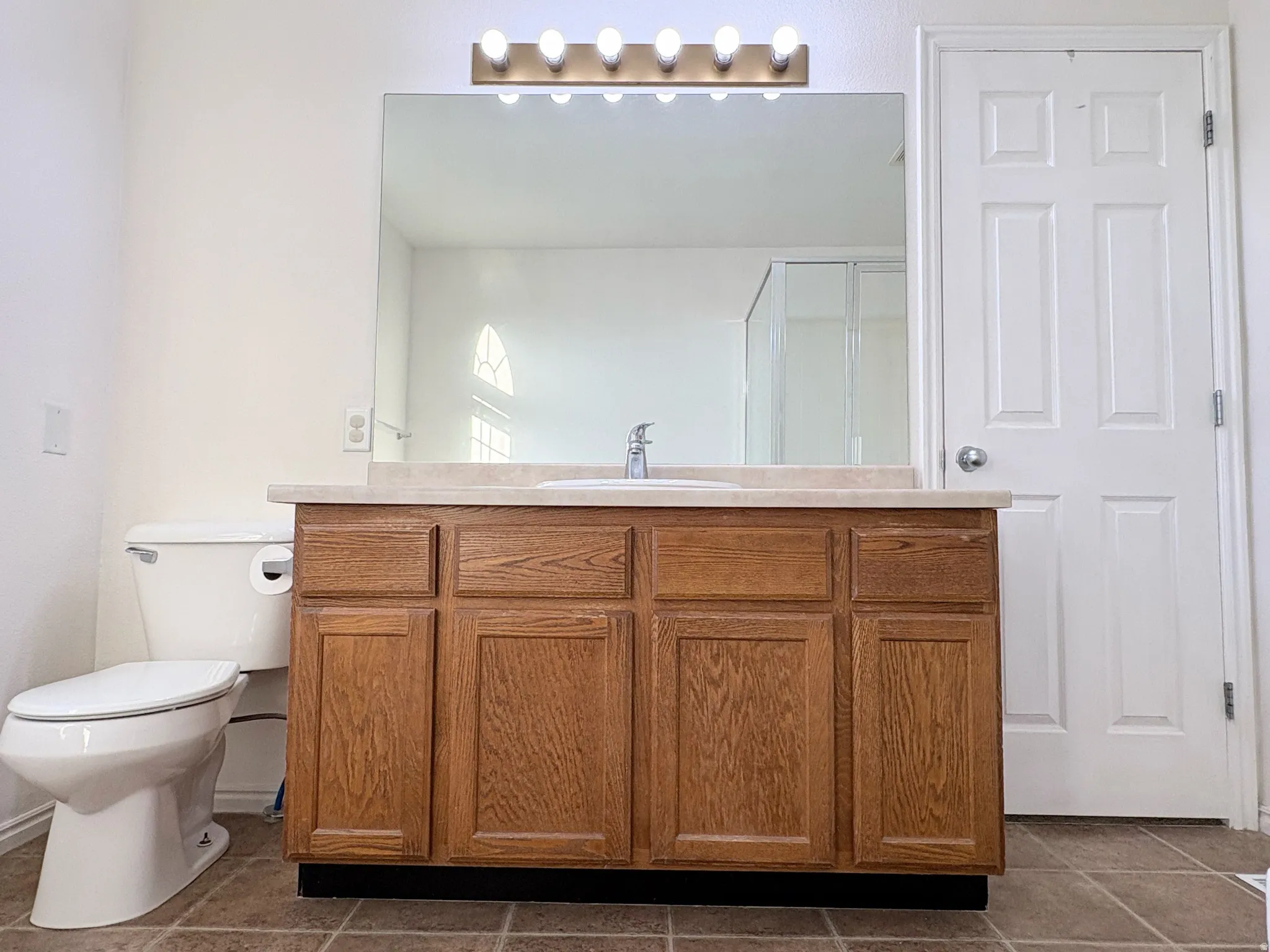 Bathroom featuring a wood-finish vanity with a single basin sink, a large wall-mounted mirror, overhead vanity lighting, and tiled flooring