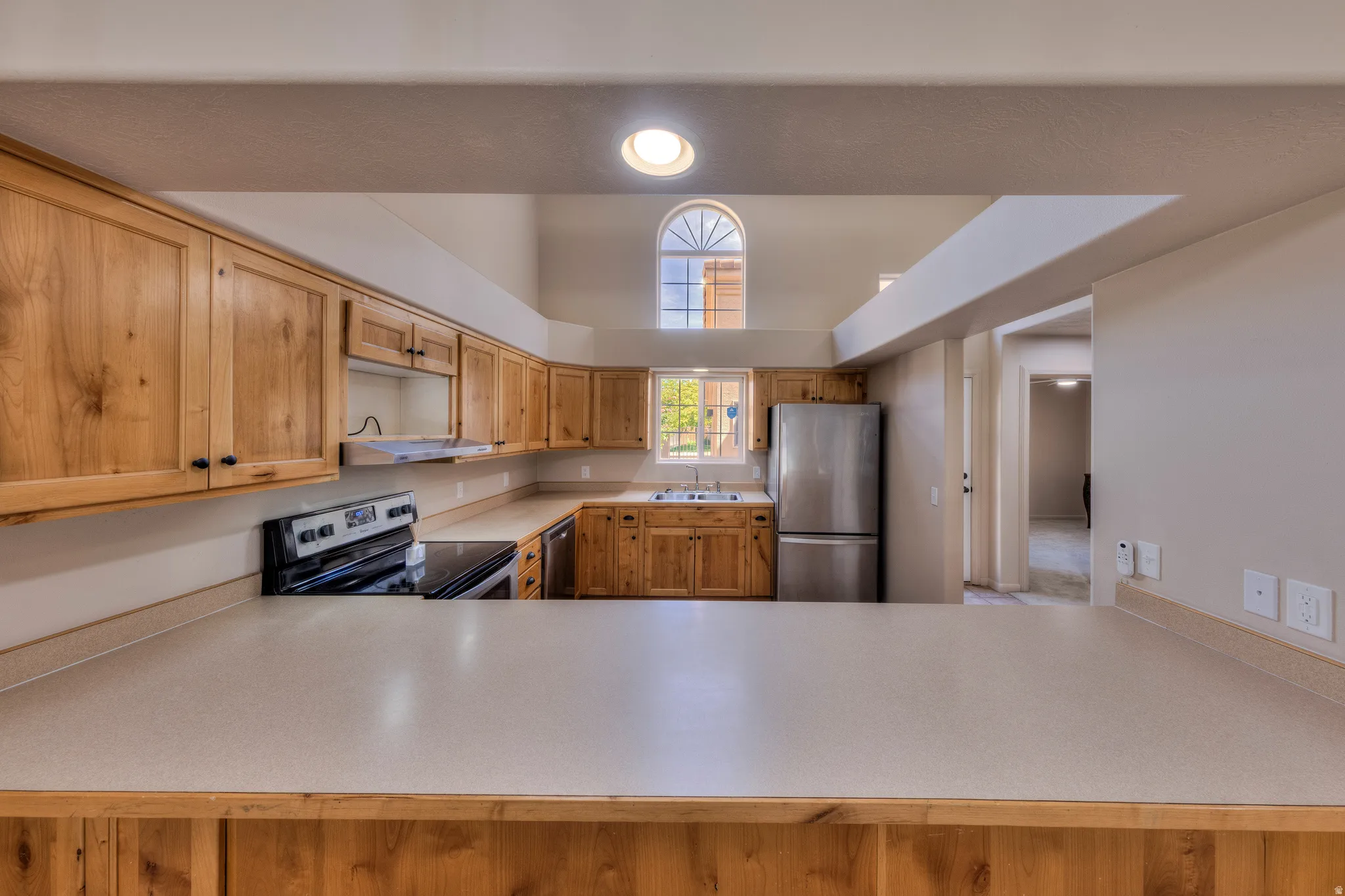 Kitchen featuring extensive wood cabinetry, light-toned countertops, stainless steel appliances, and a vaulted ceiling with an arched window