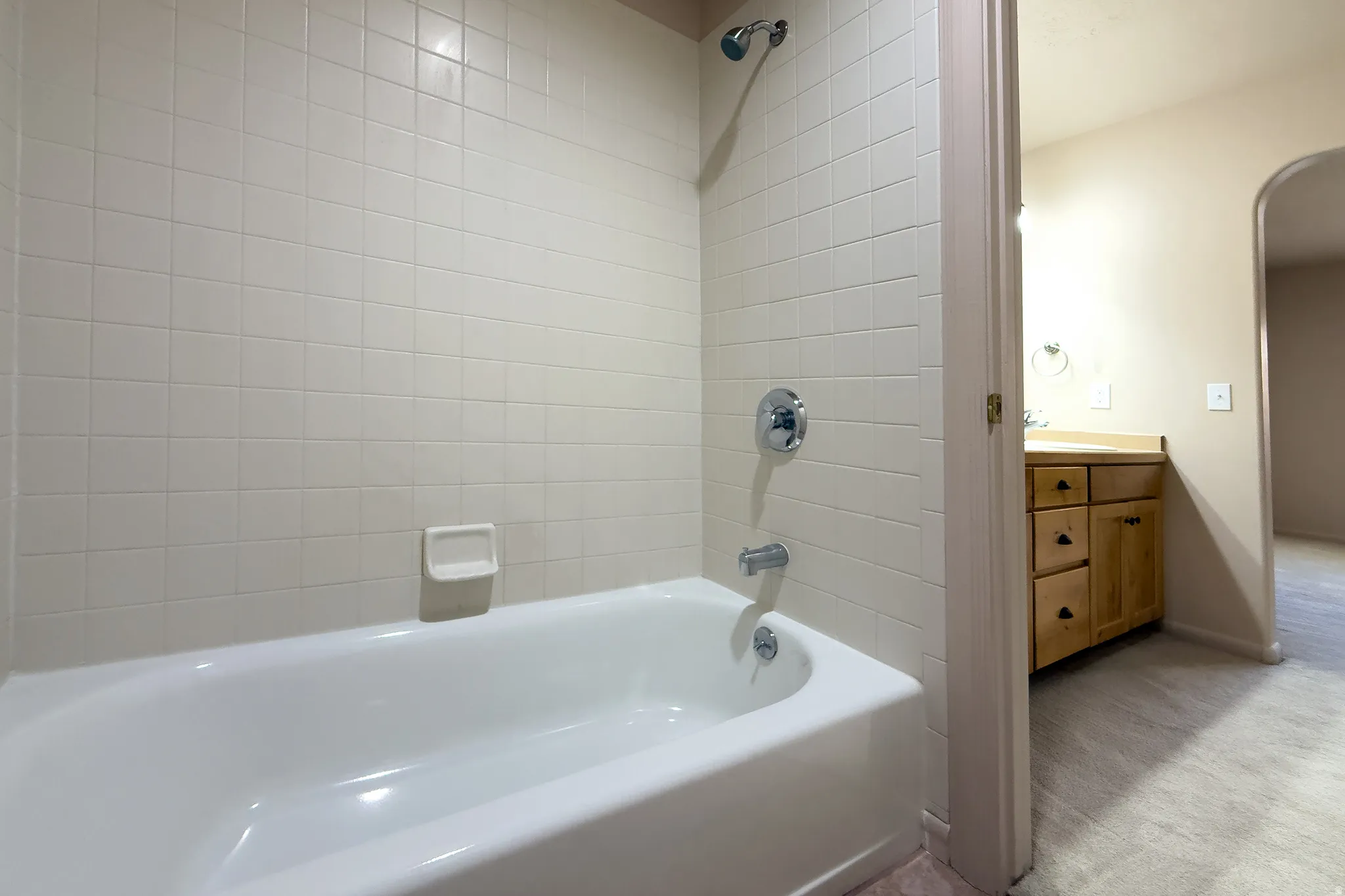 White subway tile surround with chrome fixtures, a classic white bathtub, a natural wood-finish vanity with cabinet storage and drawers, and an arched hallway opening