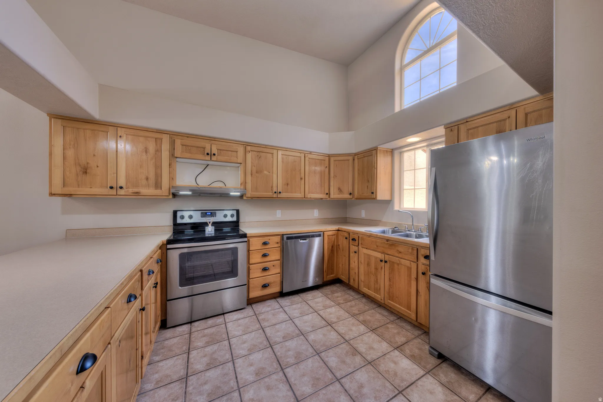 Kitchen featuring vaulted ceilings, an arched top window, light wood cabinetry, neutral countertops, and stainless-steel appliances