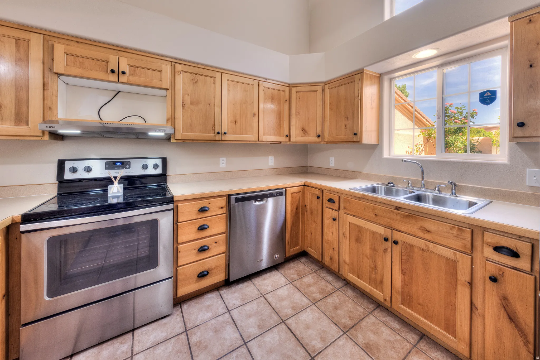 Kitchen featuring natural wood cabinetry, stainless steel appliances including a range and dishwasher, a double basin sink, and tile flooring