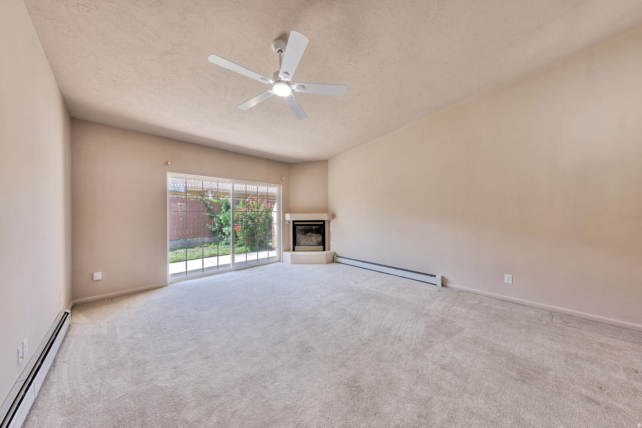 Spacious carpeted room featuring a ceiling fan, a corner fireplace with a hearth, and a sliding glass door leading to a fenced outdoor area