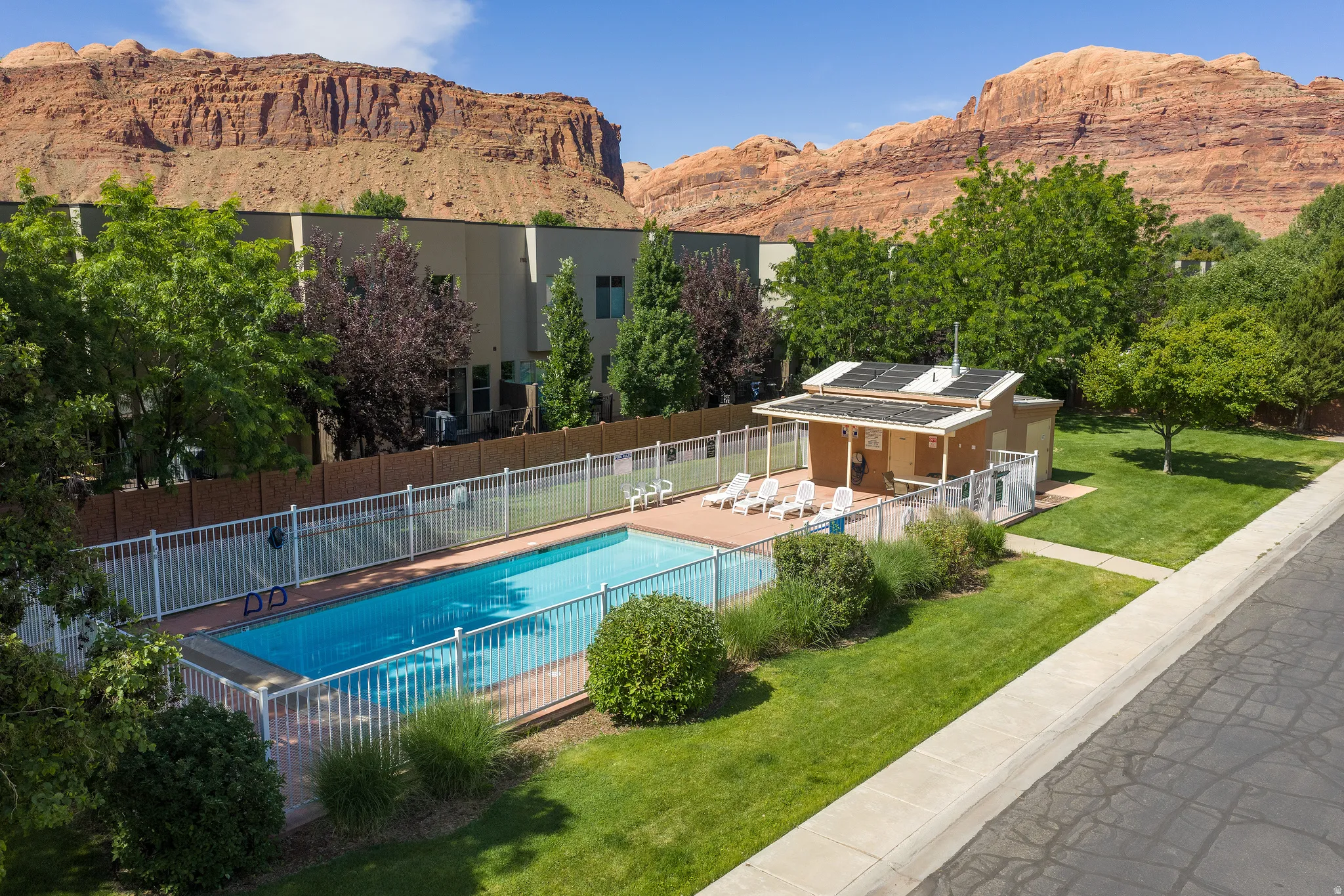 Fenced swimming pool area featuring a pool house with solar panels, surrounded by a manicured lawn and mature trees