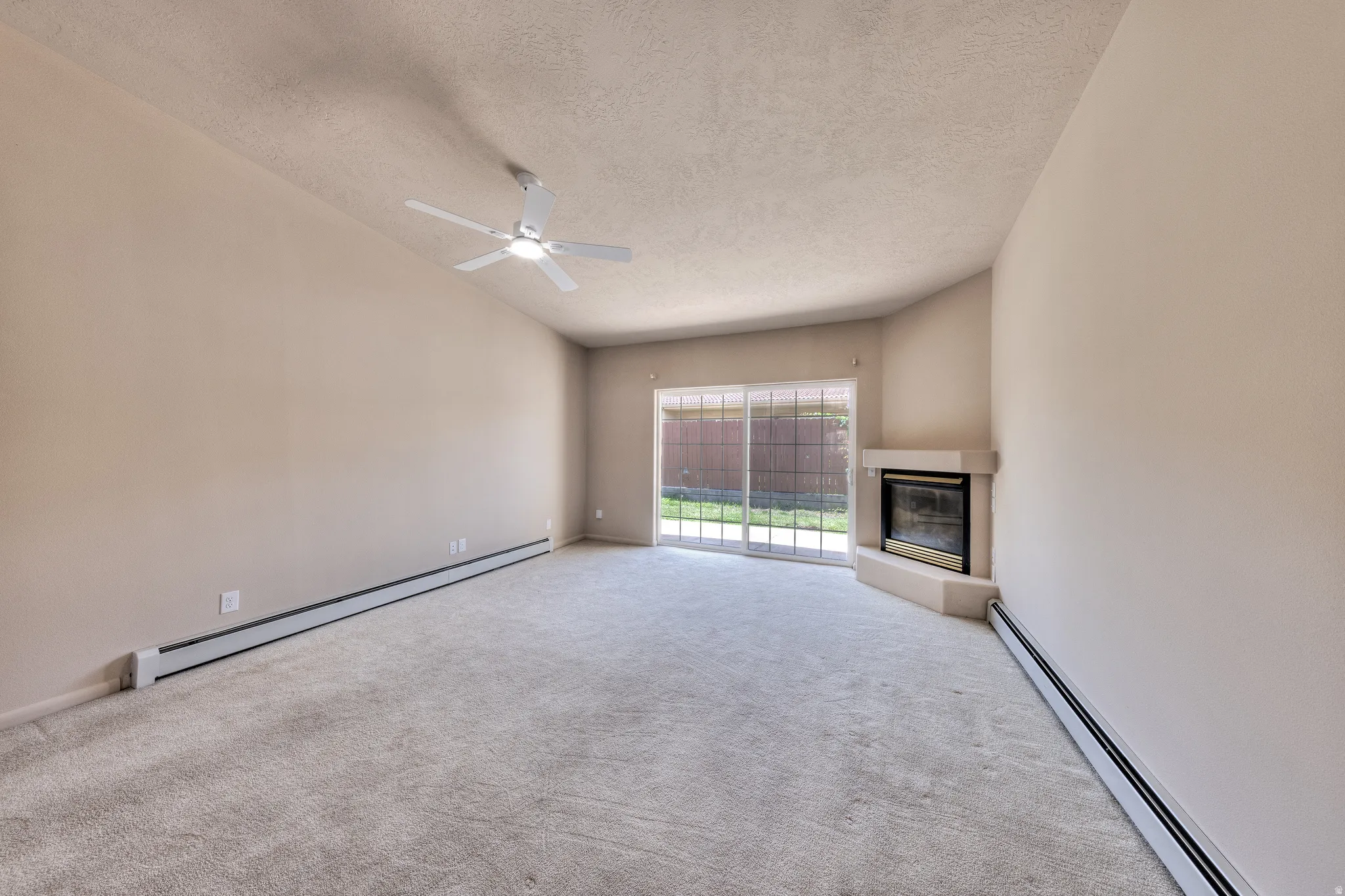 Carpeted living area featuring a corner fireplace with a light-toned surround, a ceiling fan, and sliding glass doors opening to a fenced outdoor area with grass
