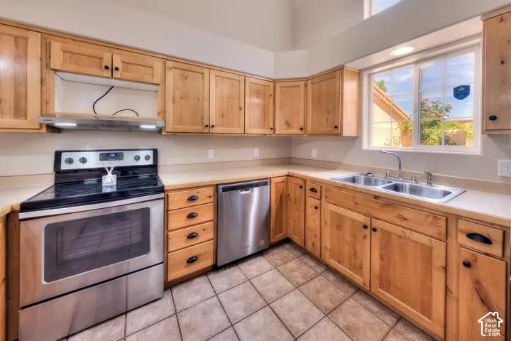 Kitchen featuring natural wood cabinetry, stainless steel appliances including a range and dishwasher, a double basin sink, and tile flooring