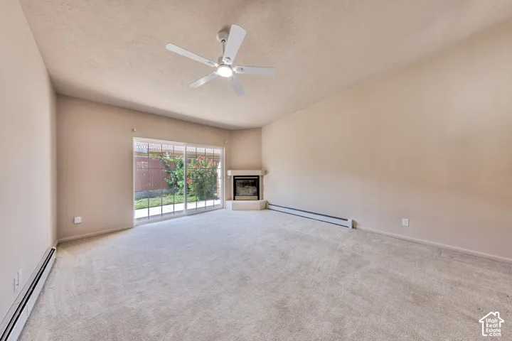 Spacious carpeted room featuring a ceiling fan, a corner fireplace with a hearth, and a sliding glass door leading to a fenced outdoor area
