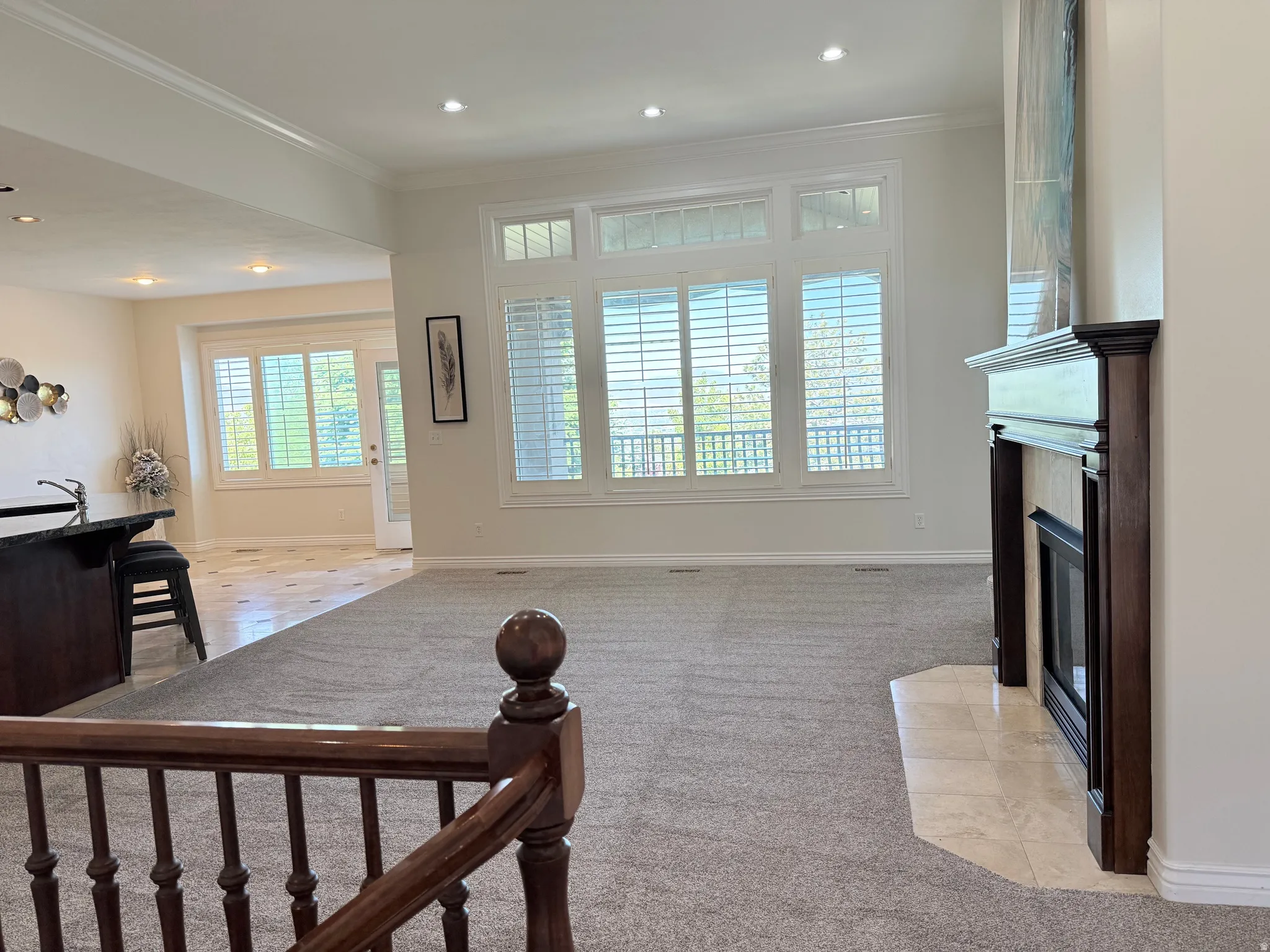 Open-concept living space with light gray carpeting, a dark wood-finish fireplace surround with a tiled hearth, and large windows featuring plantation shutters