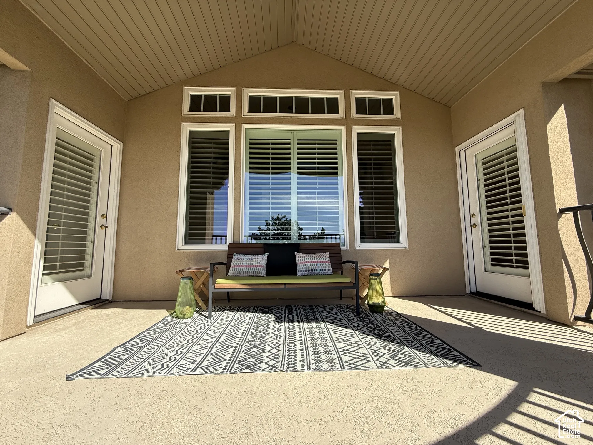 Covered patio featuring an angled wood-finish ceiling, textured exterior walls, multiple windows with plantation shutters, and two glass-paneled doors with internal blinds