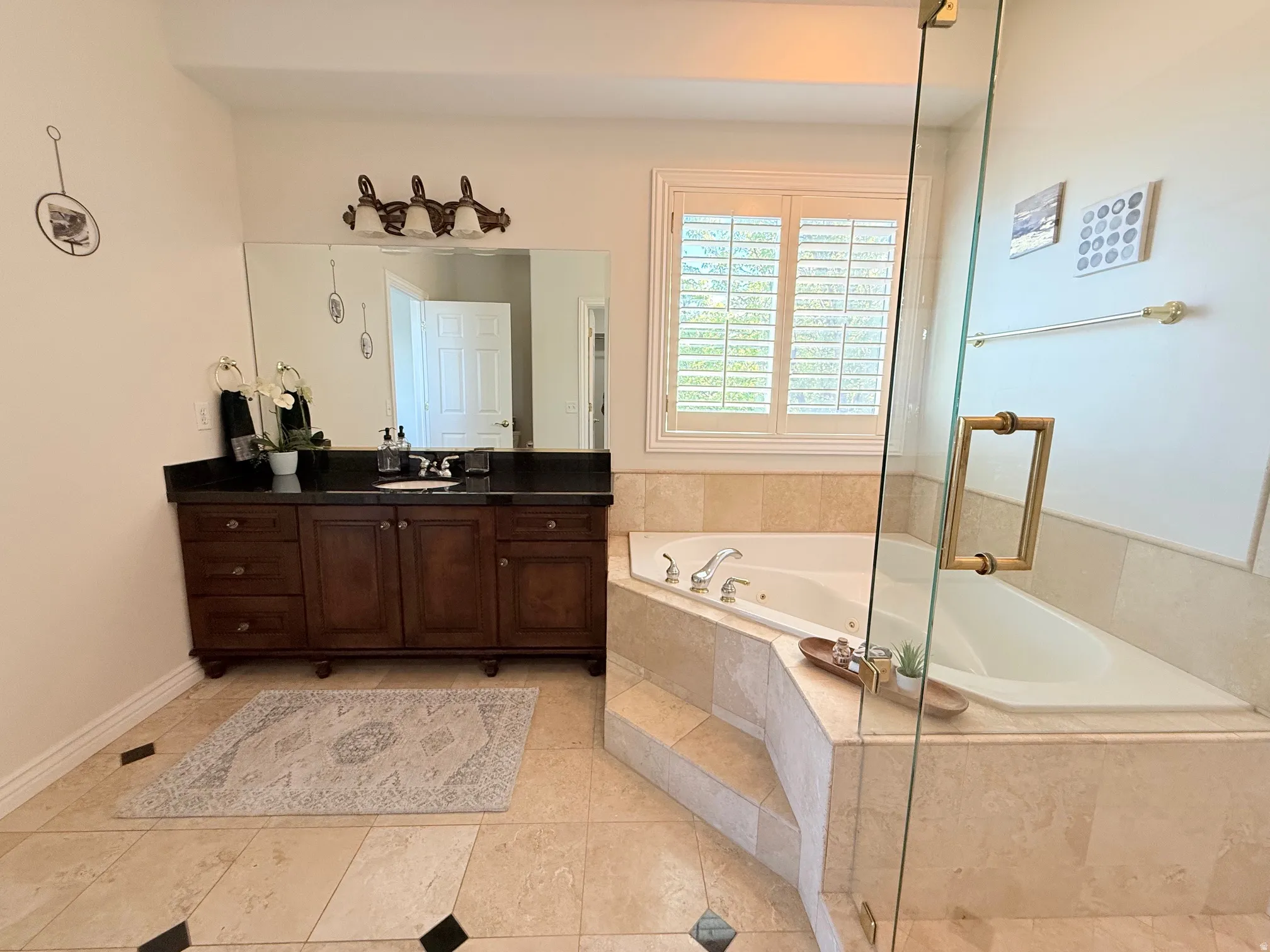 Built-in corner soaking tub with surround, dark wood vanity with a black countertop, large vanity mirror, plantation shutters, and glass shower enclosure with brass-tone hardware
