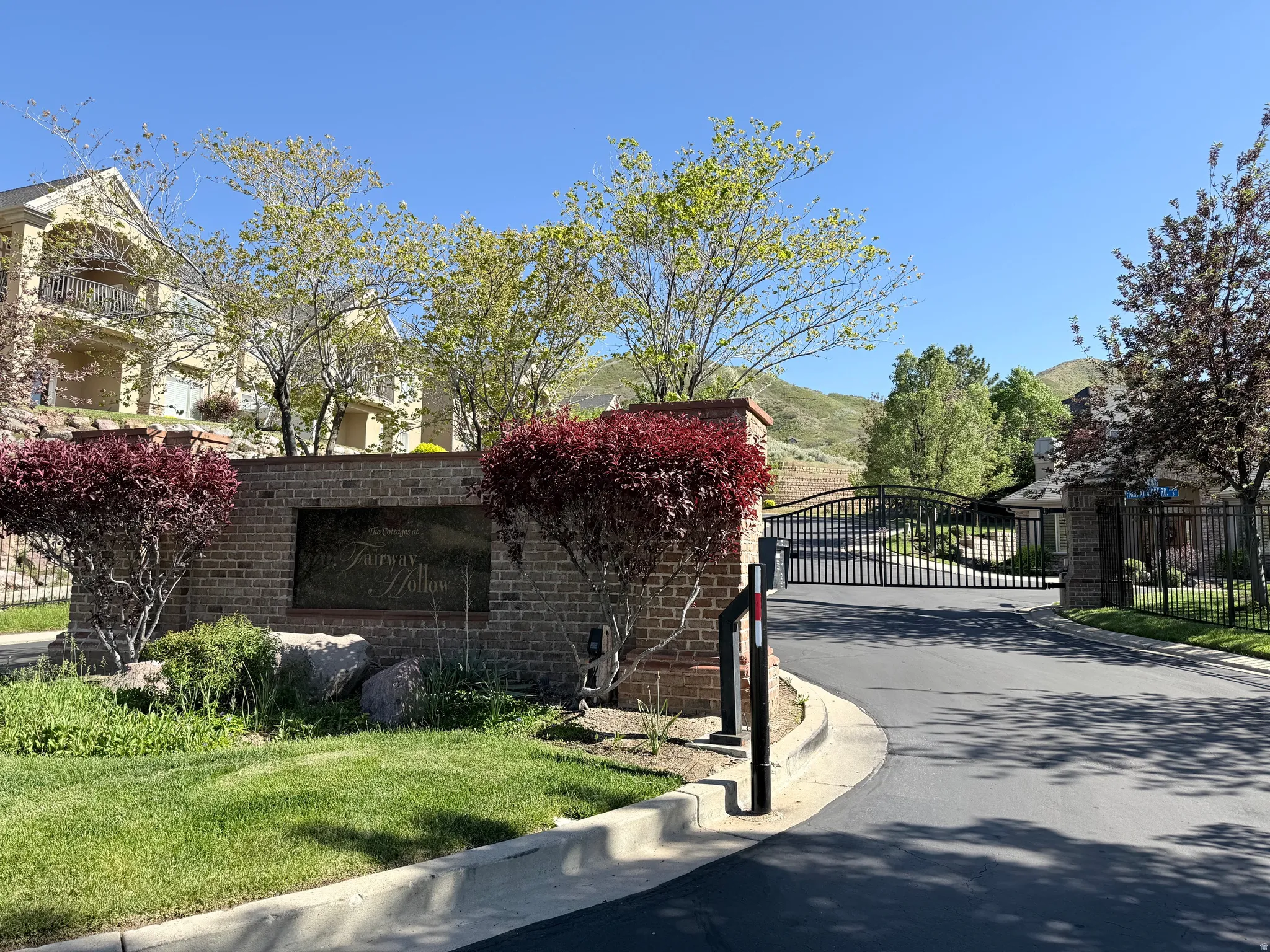 Gated community entrance featuring a brick monument sign, wrought iron gates, mature landscaping, and an asphalt driveway