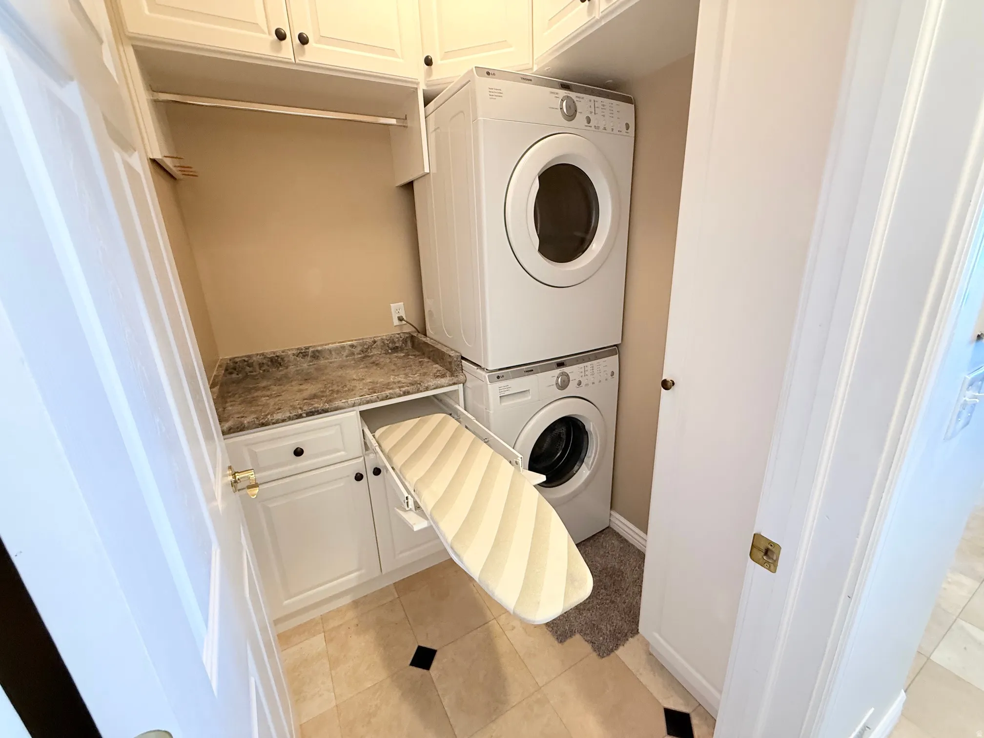 Dedicated laundry area featuring stacked appliances, overhead cabinetry, a countertop with a built-in ironing board, and tile flooring