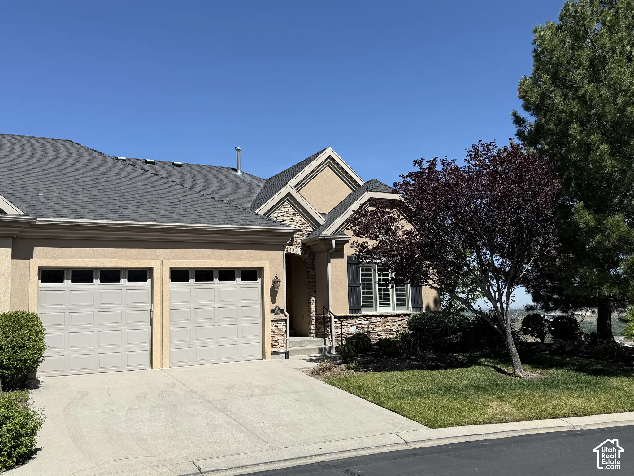 Stucco and stone exterior featuring a two-car garage, dark asphalt shingle roof, and a mature tree providing shade