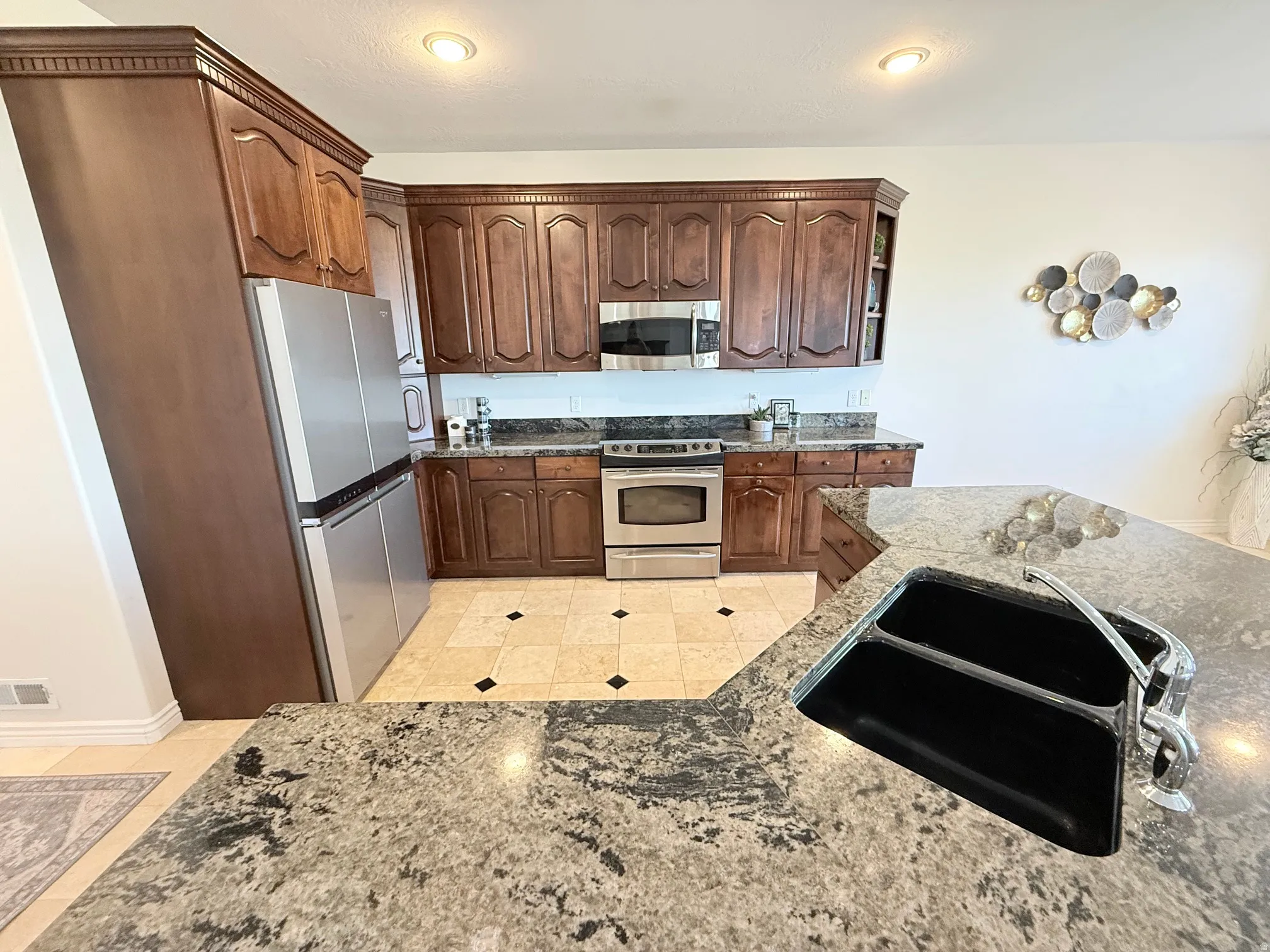 Kitchen featuring a large central island with a double basin sink, dark wood cabinetry with crown molding, stainless steel appliances, and light-toned tile flooring with dark accent tiles
