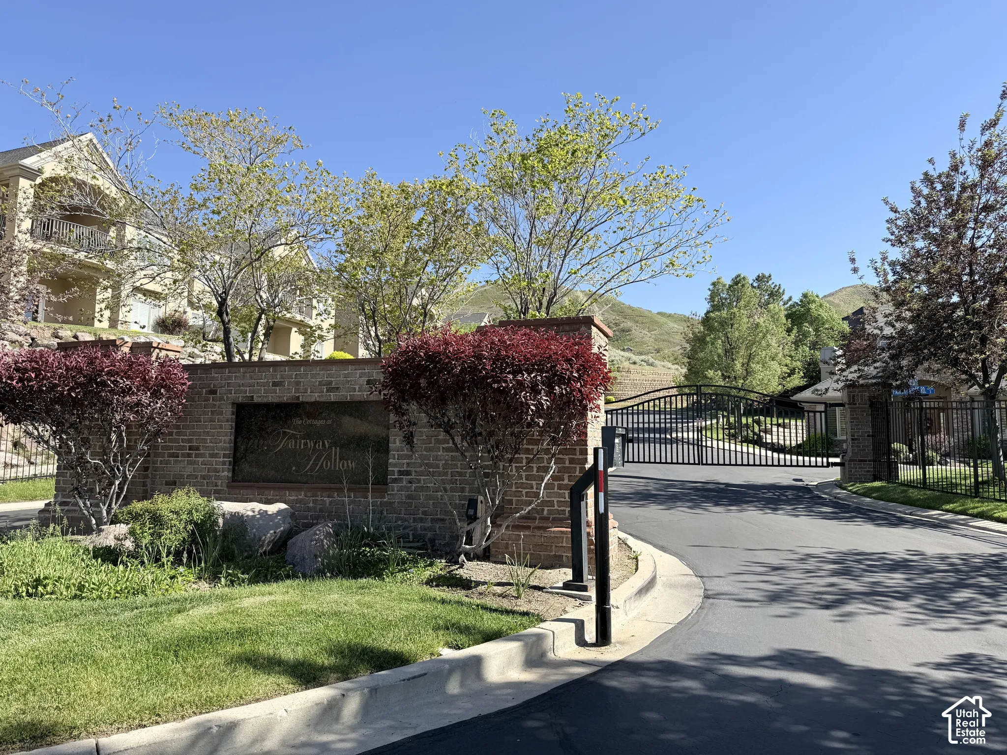 Gated community entrance featuring a brick monument sign, wrought iron gates, mature landscaping, and an asphalt driveway