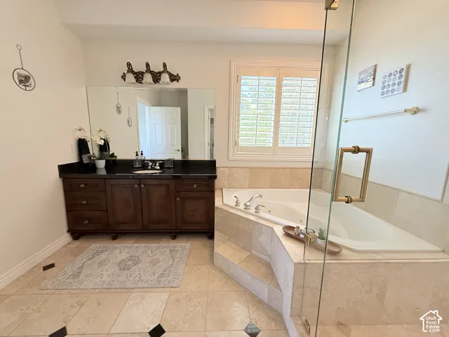Built-in corner soaking tub with surround, dark wood vanity with a black countertop, large vanity mirror, plantation shutters, and glass shower enclosure with brass-tone hardware