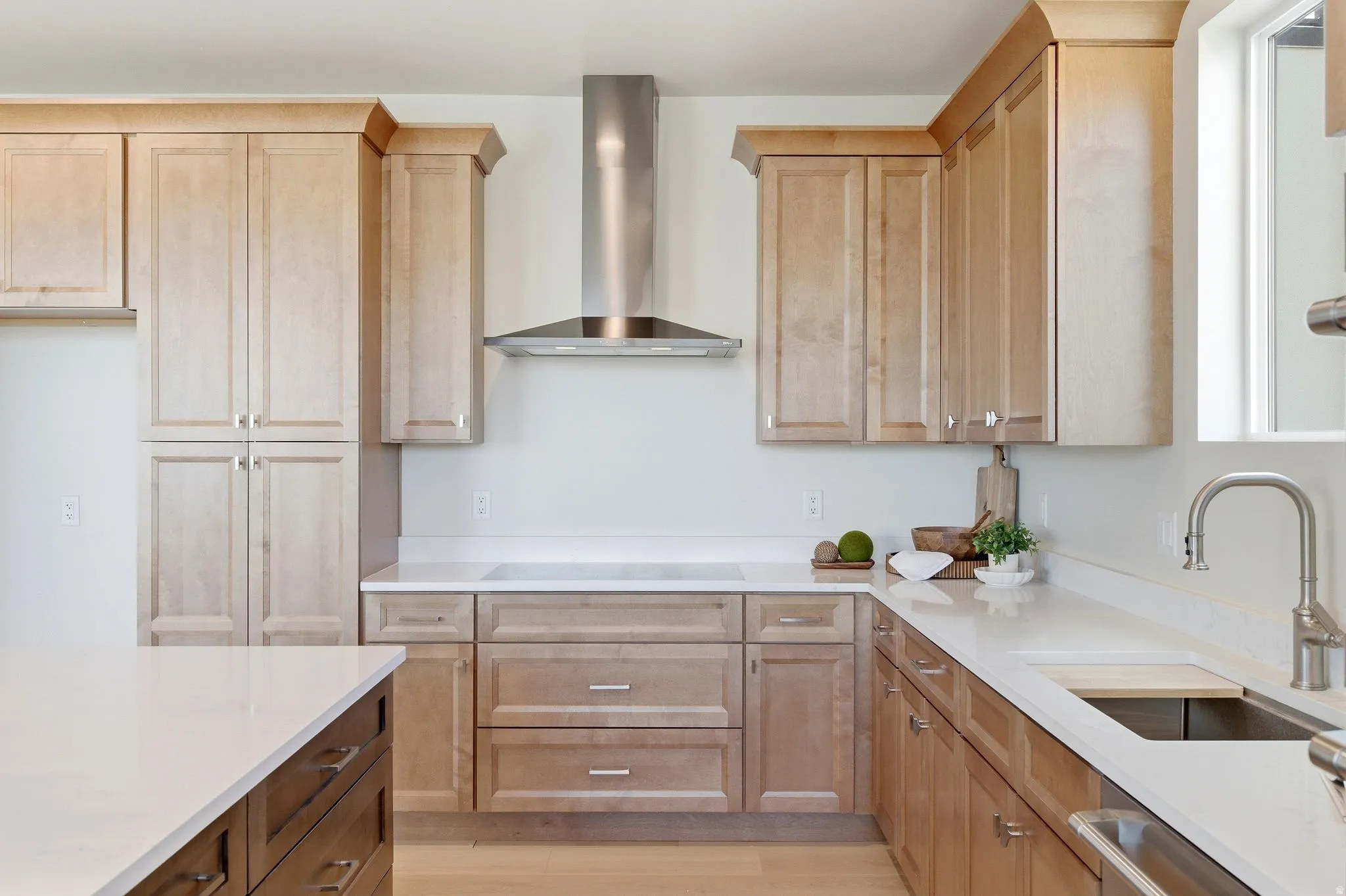Modern kitchen featuring light wood-finish cabinetry, stainless steel range hood, white countertops, built-in cooktop, and a stainless steel sink with a gooseneck faucet