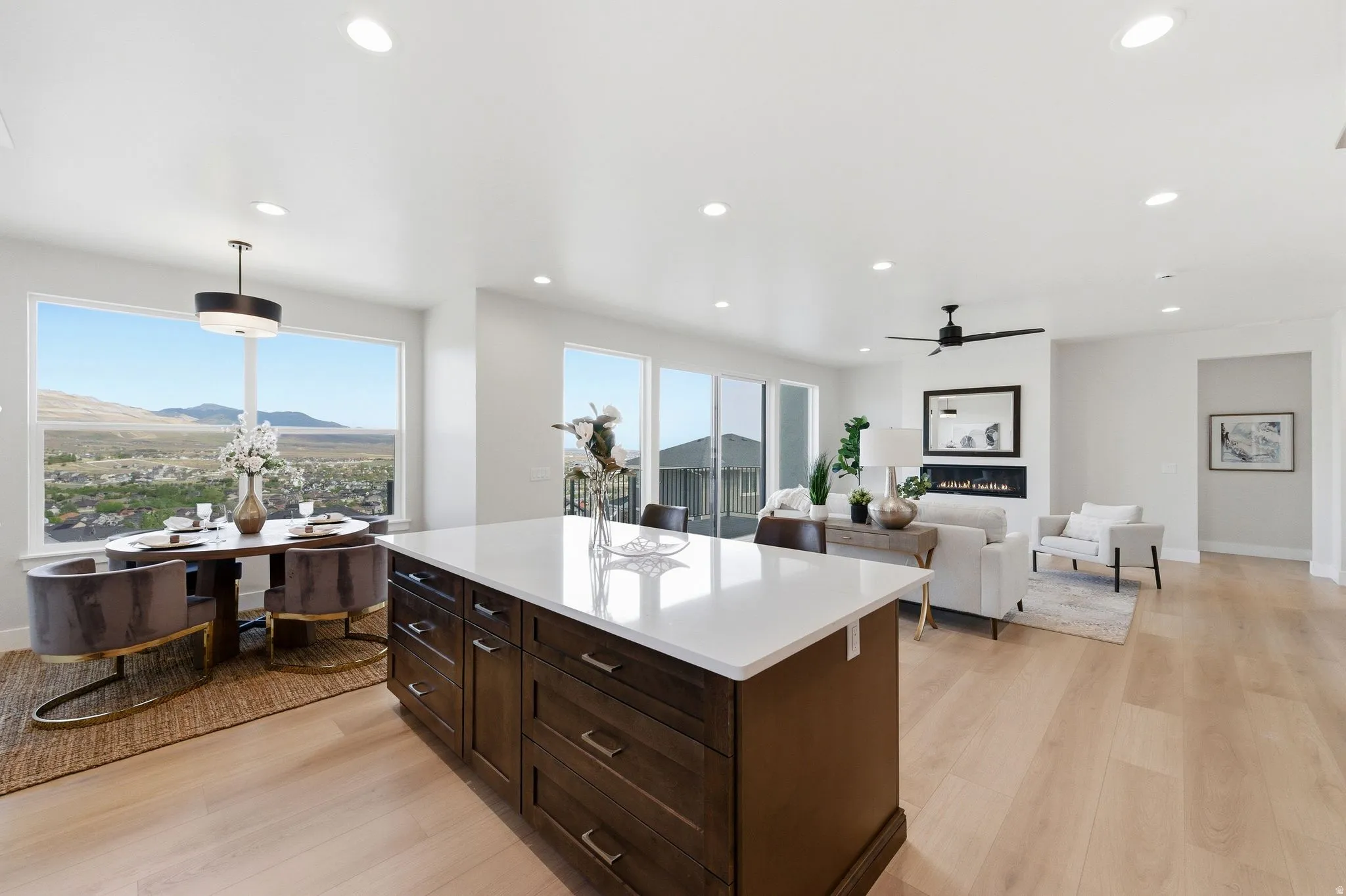Open-concept living area featuring a large kitchen island with white countertop, light wood-finish flooring, recessed lighting, a built-in electric fireplace, and expansive windows