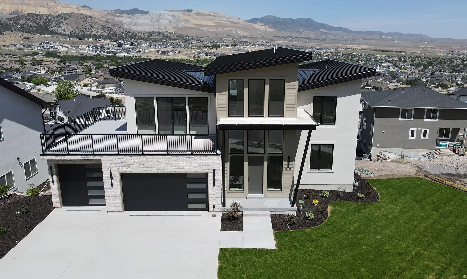 Modern architectural exterior featuring a prominent balcony, contemporary garage doors, stucco and stone-finish siding, a concrete driveway, and landscaped front yard