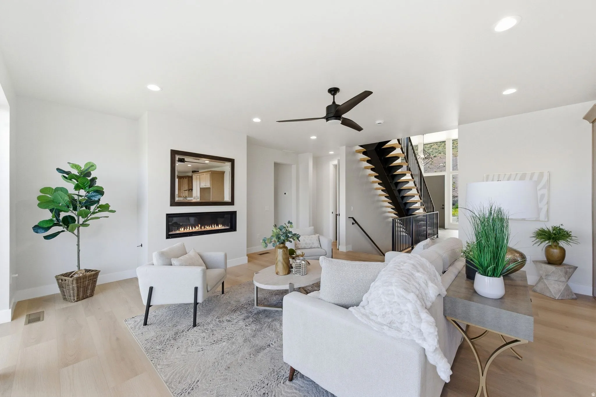 Spacious living area featuring light wood-finish flooring, a linear electric fireplace with a dark-framed mirror, a contemporary ceiling fan, recessed lighting, and an open-tread staircase with dark risers and railings