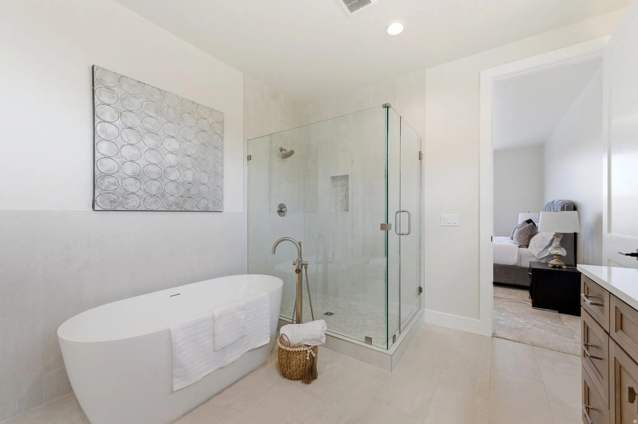 Spacious bathroom featuring a freestanding soaking tub, a glass-enclosed shower with built-in niche, light-toned wall tile, wood-finish vanity with countertop, and recessed lighting