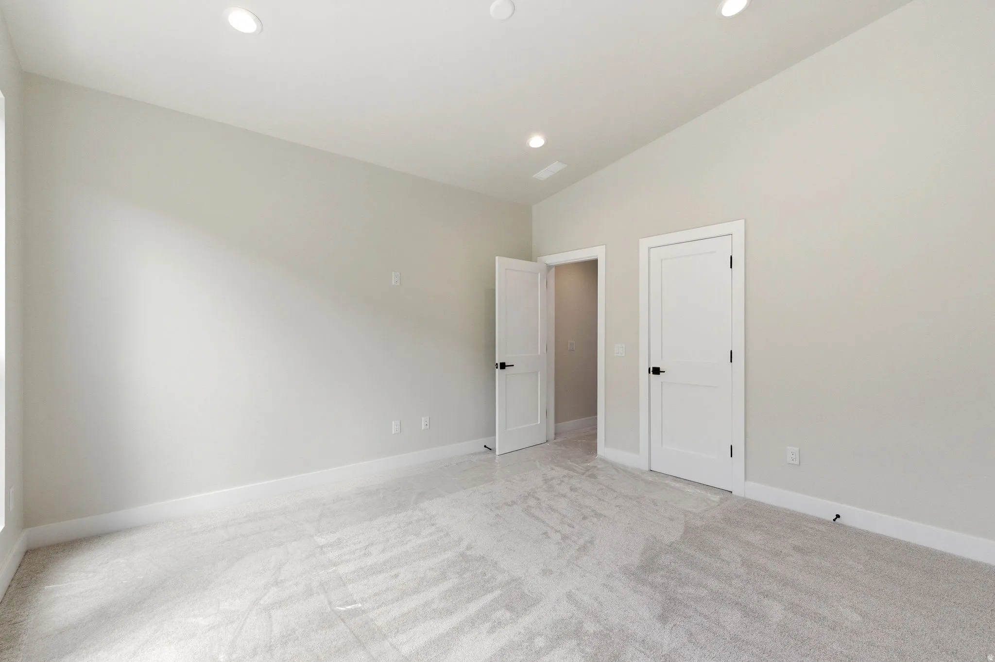 Spacious room featuring light gray carpeting, recessed ceiling lighting, two white panel doors with black hardware, and crisp white baseboards