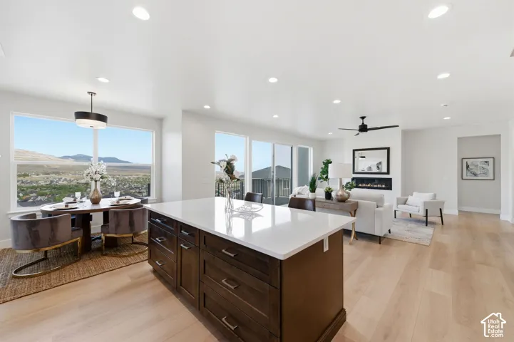 Open-concept living area featuring a large kitchen island with white countertop, light wood-finish flooring, recessed lighting, a built-in electric fireplace, and expansive windows