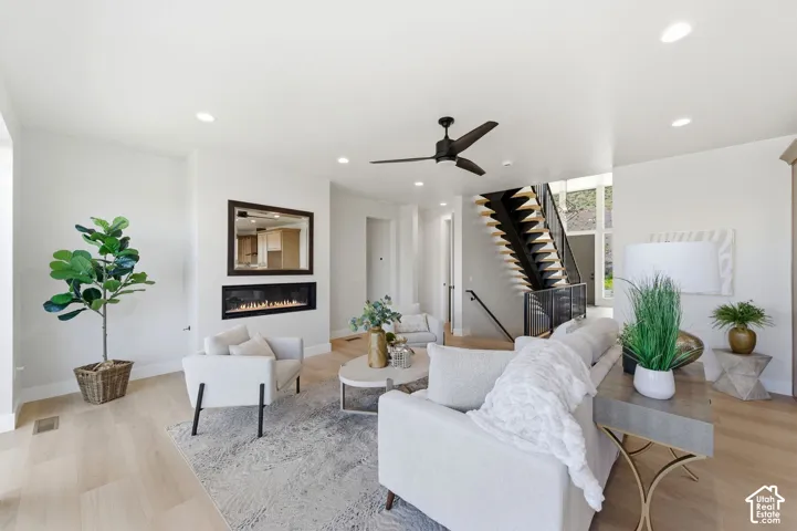 Spacious living area featuring light wood-finish flooring, a linear electric fireplace with a dark-framed mirror, a contemporary ceiling fan, recessed lighting, and an open-tread staircase with dark risers and railings