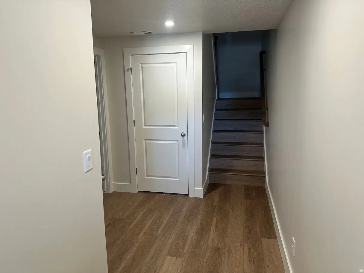 Hallway featuring wood-finish flooring, white two-panel door with brushed nickel hardware, light neutral wall paint, white baseboards, and recessed ceiling lighting