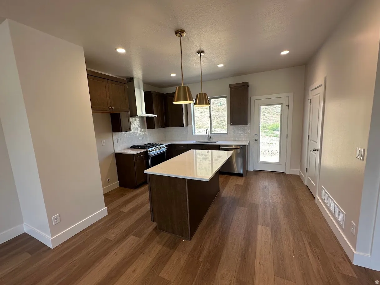 Contemporary kitchen featuring wood-finish flooring, dark wood cabinetry, stainless steel appliances, a white tile backsplash, and a central island with a light-toned countertop