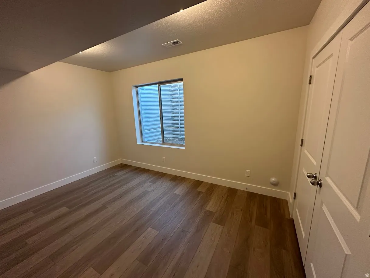 Room featuring wood-finish flooring, a window with a metal window well, light-toned wall paint, and a double-door closet with brushed nickel hardware