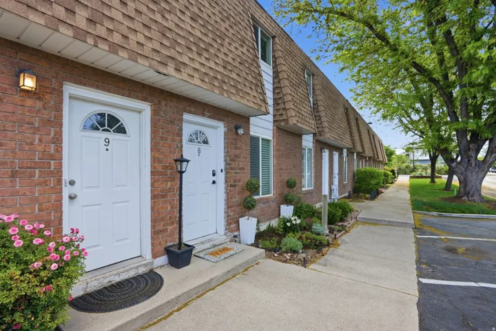Brick townhouse exterior featuring white entry doors, pitched shingle roofing, and established landscaping
