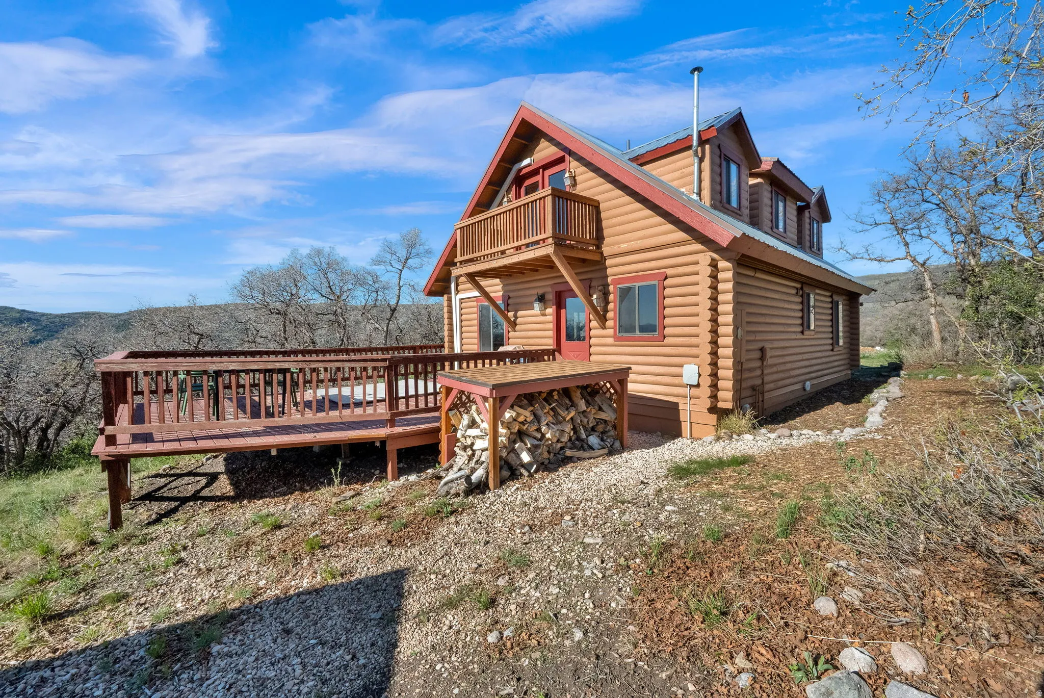 Log cabin exterior featuring a multi-level deck, a second-story balcony, and a covered wood storage area