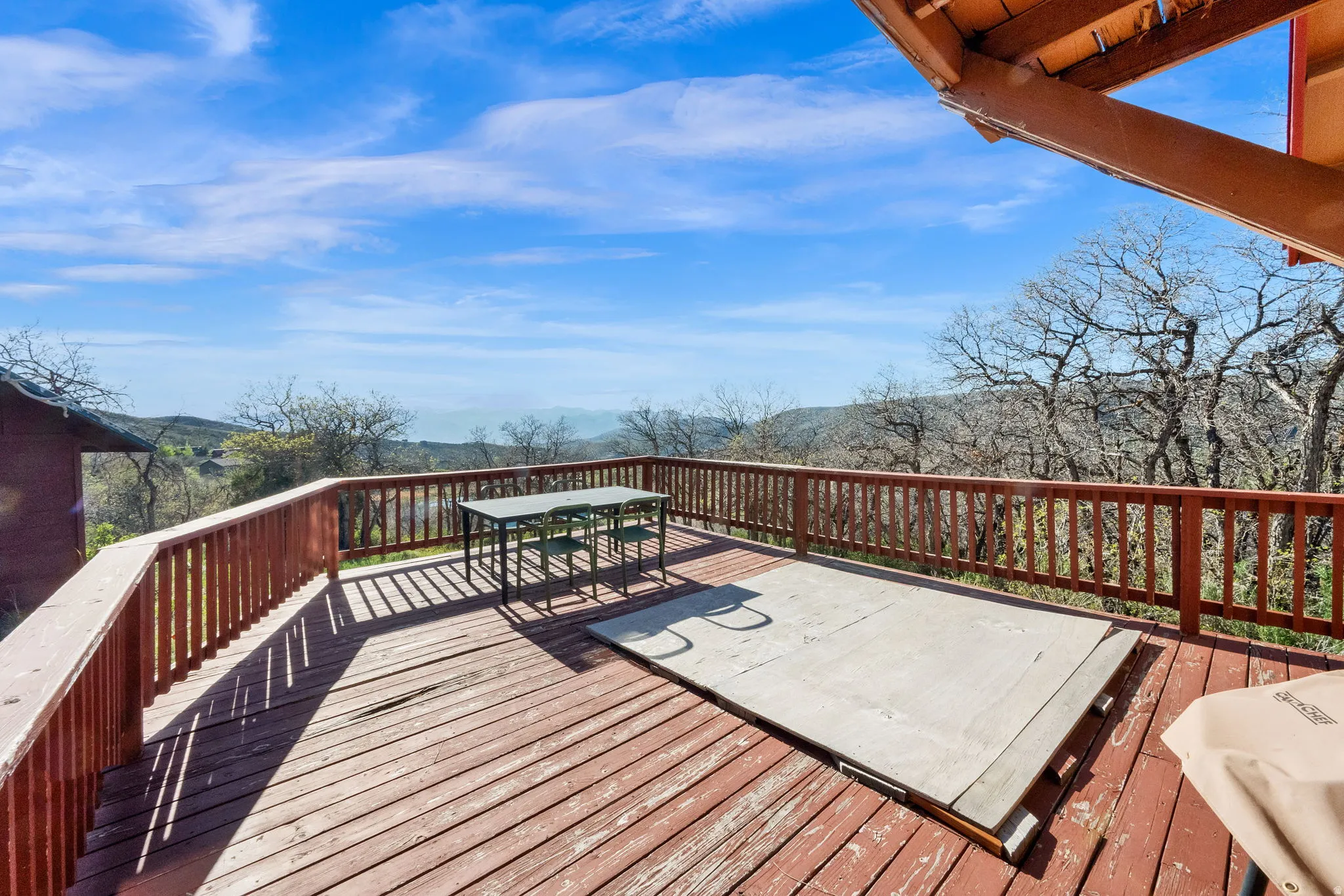 Expansive wood deck with wood railings, surrounded by mature deciduous trees and distant mountain scenery under a bright blue sky