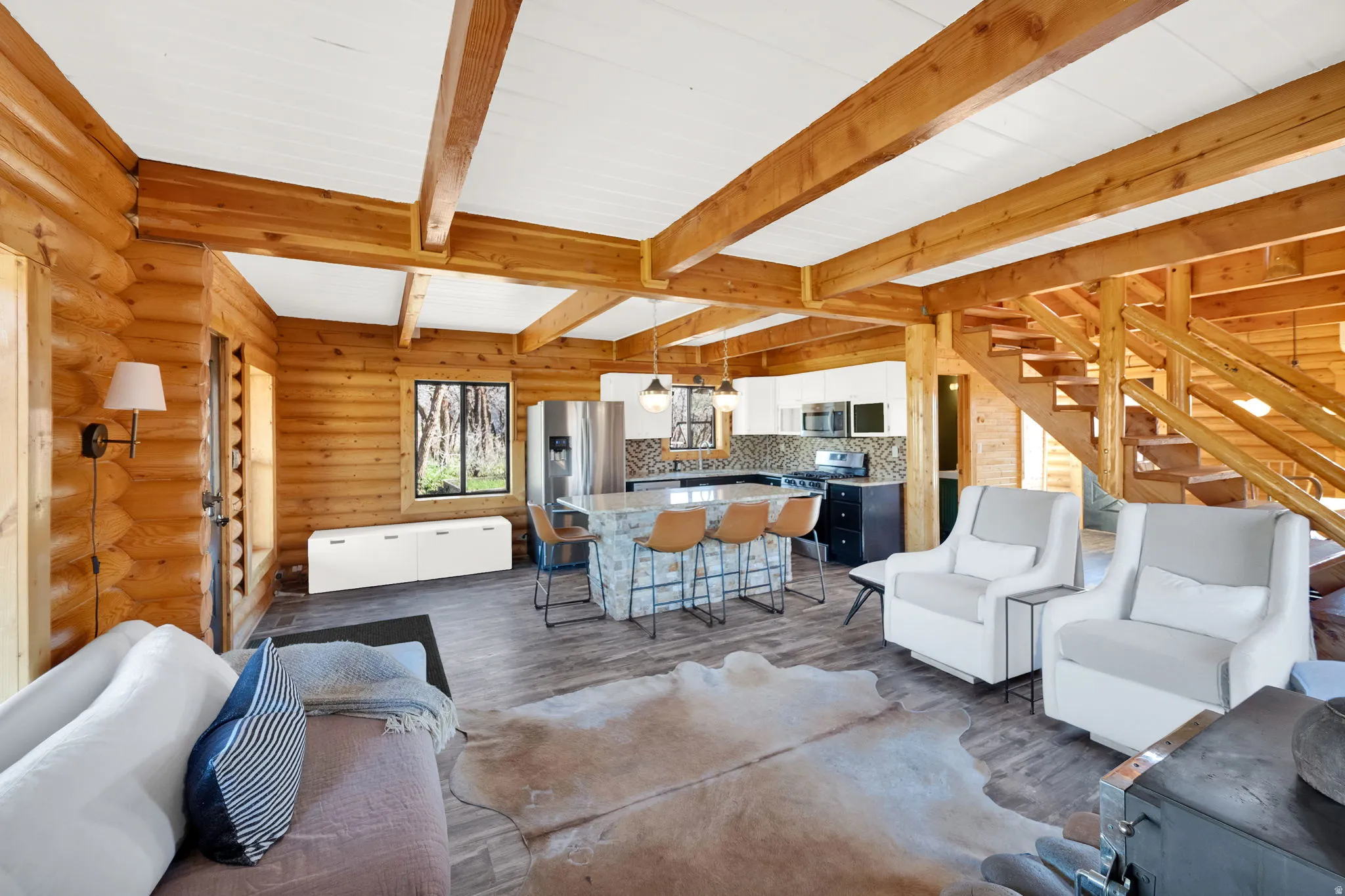 Open-concept room featuring exposed wood beams, log walls, wood-finish flooring, a kitchen island with seating, and a visible staircase