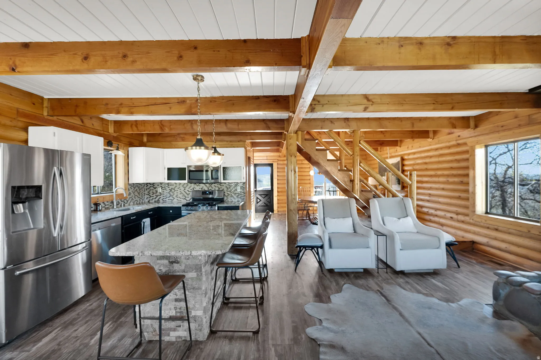 Open concept living space featuring a gourmet kitchen, exposed wood beams, wood-finish flooring, a log cabin wall, and a staircase with natural wood railings