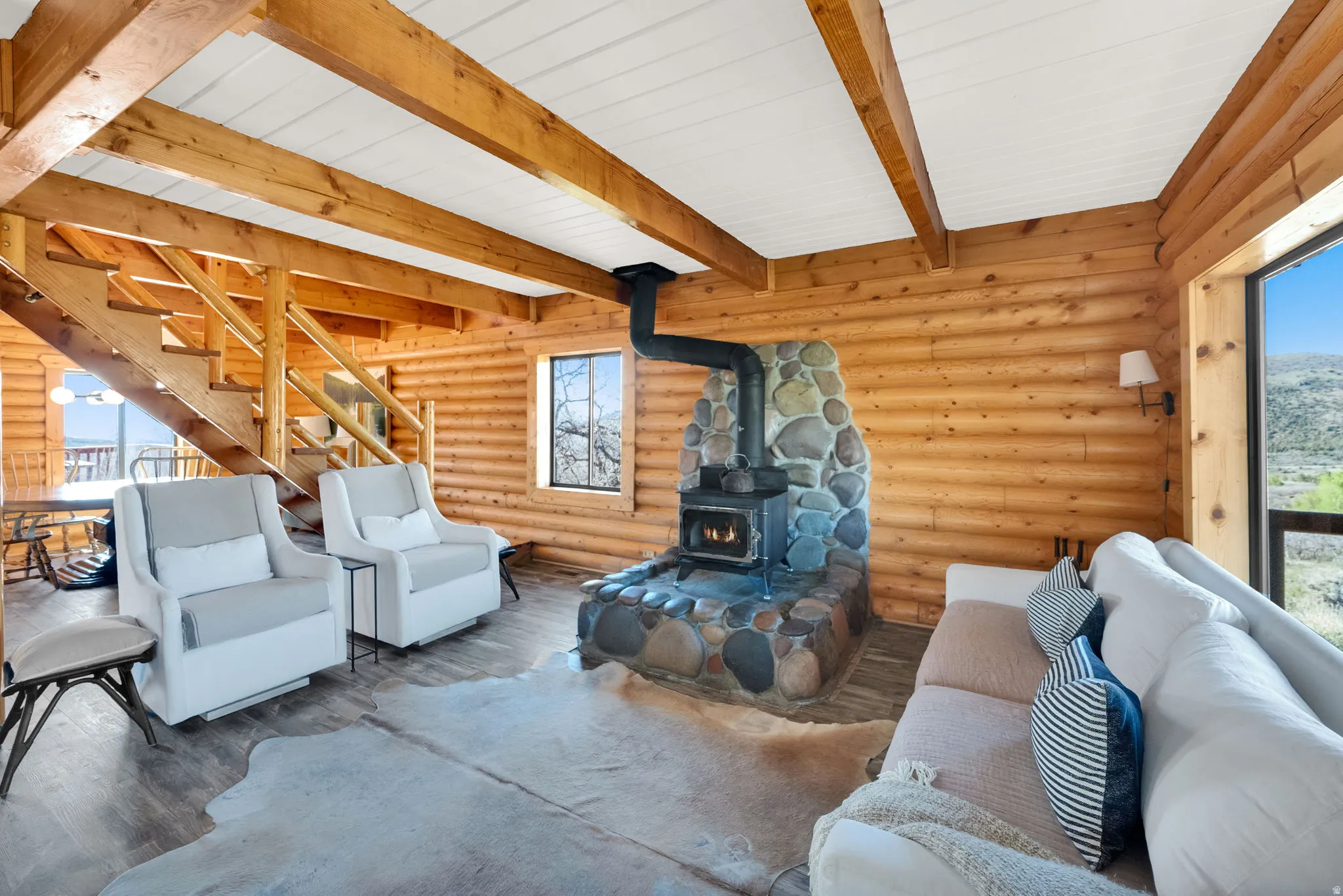 Log cabin interior featuring a stone-faced wood-burning stove, exposed wood beams, wood-finish flooring, and a wooden staircase