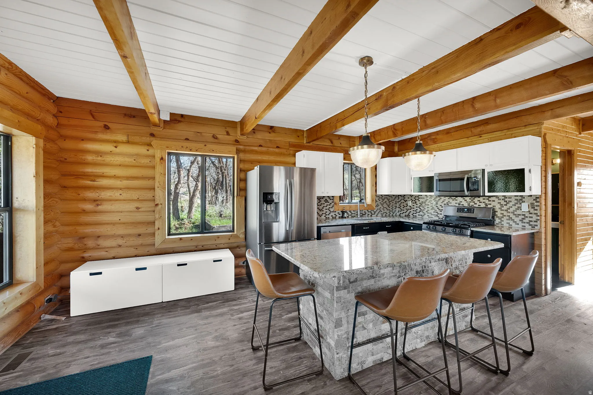 Spacious kitchen featuring exposed wood beams, a large island with stone veneer and a light-colored countertop, stainless steel appliances, and wood-finish flooring