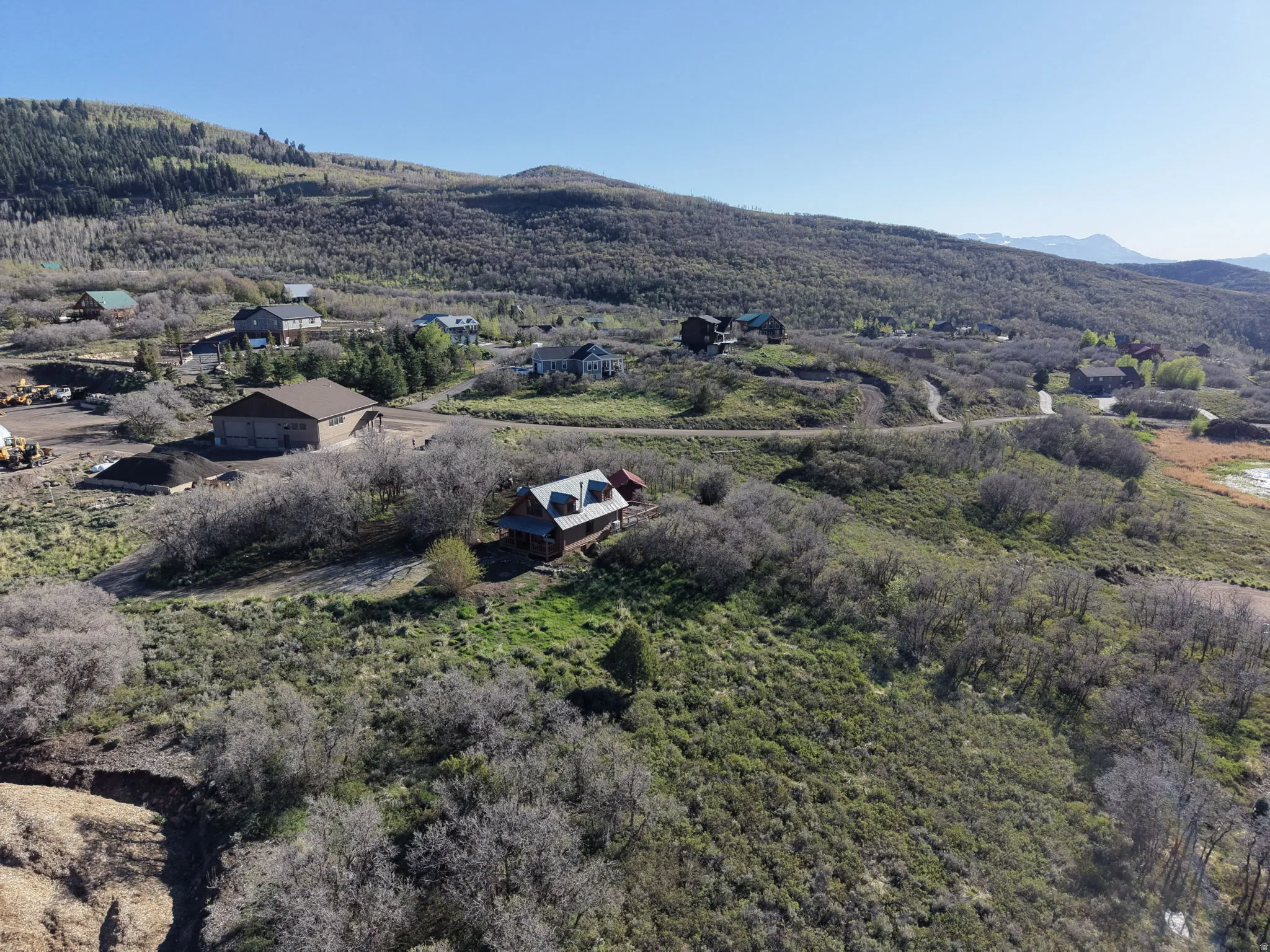 Hillside property featuring a rustic-style cabin with a red roof, surrounded by extensive natural vegetation, a winding road, and distant mountain ranges