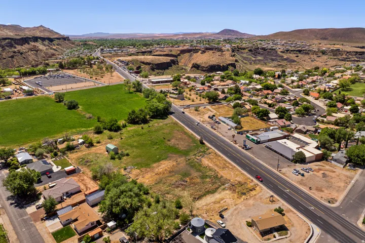 Expansive undeveloped land parcels alongside a paved road, featuring surrounding residential and commercial properties, a manicured green field, and distant elevated terrain